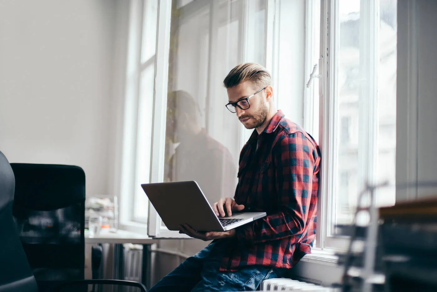 A man wearing a red and blue flannel shirt uses his laptop while he leans on a wall.