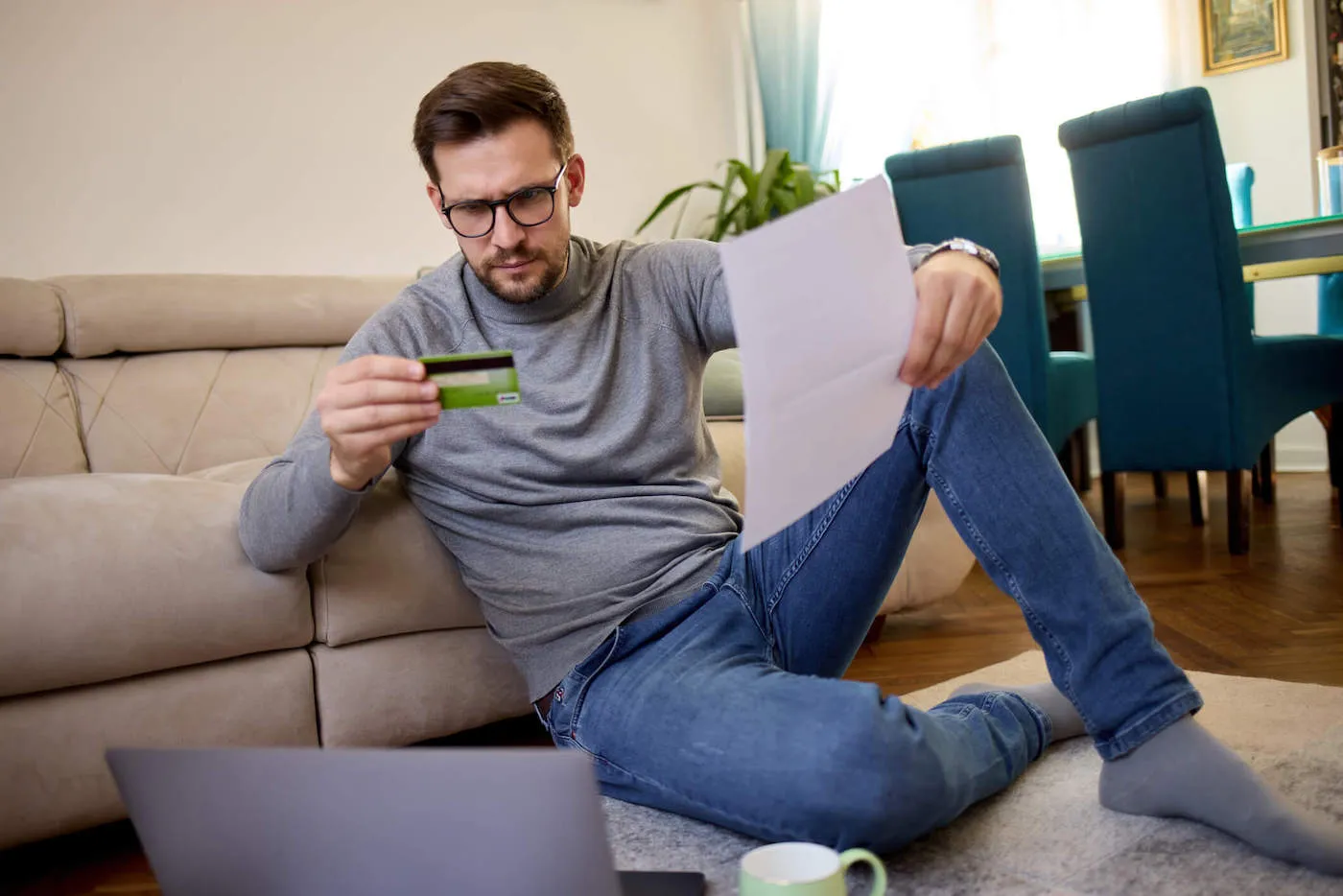 A man wearing a gray shirt and glasses frowns while holding a credit card and a document.