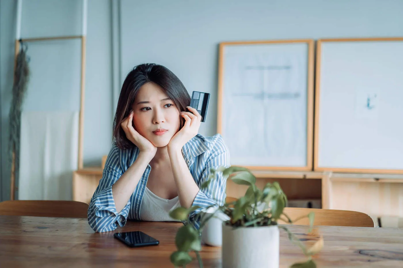 A woman frowns as she hold her hands to her face while holding her credit card with her phone on the table.