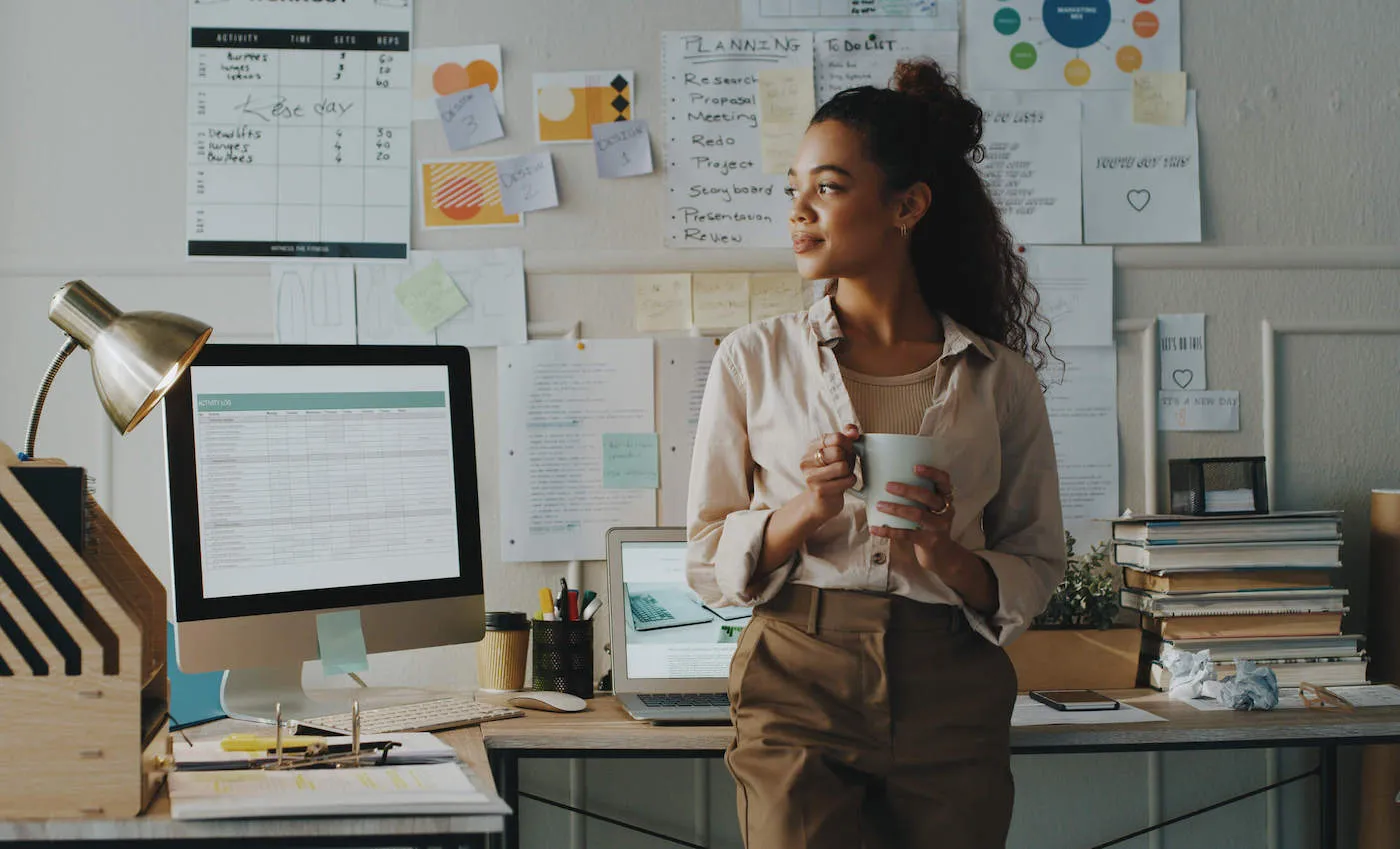 A woman holding a coffee cup smiles inside her office with documents attached to the wall in the background.