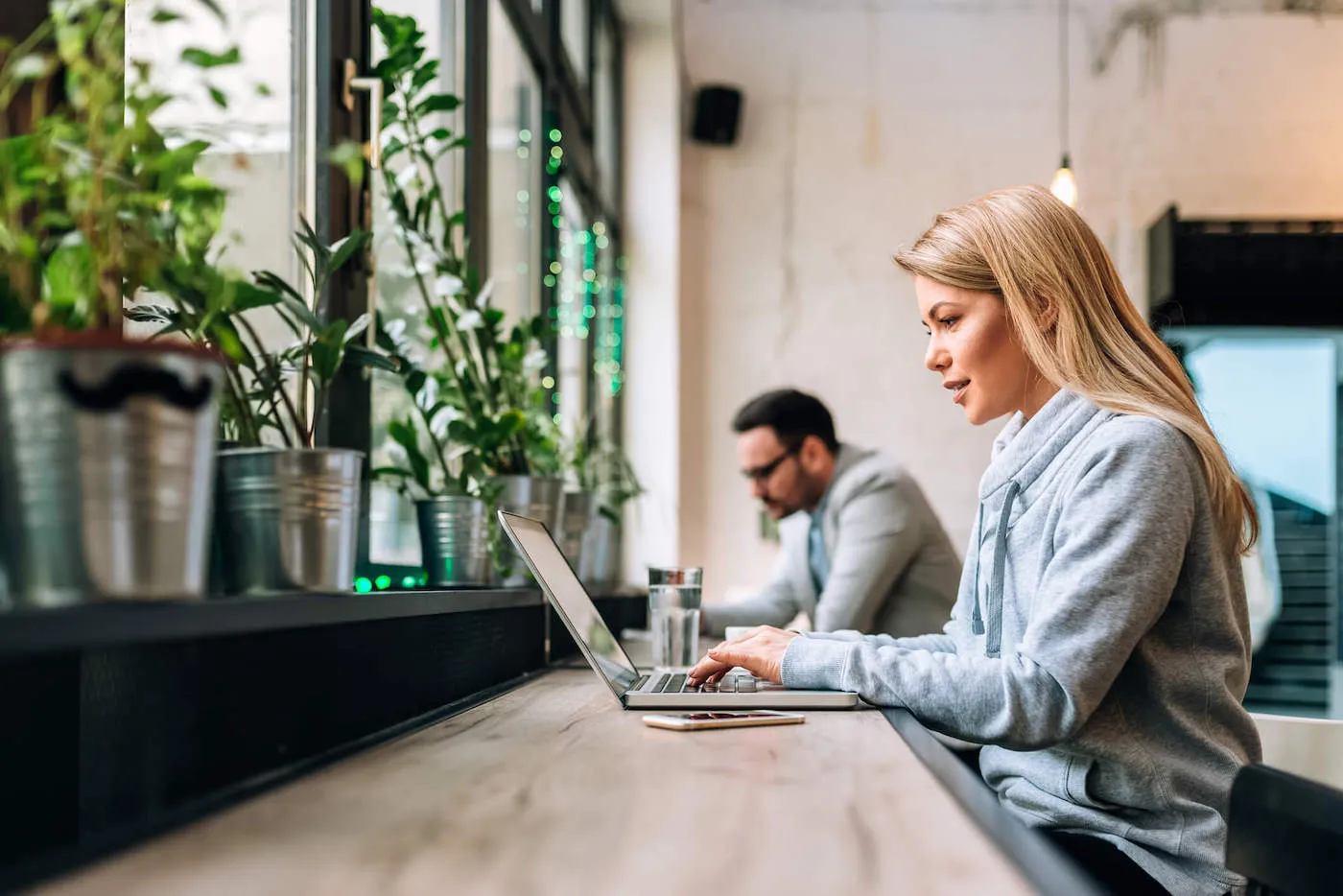 A woman wearing a gray sweater uses her laptop while potted plants are in front of her.