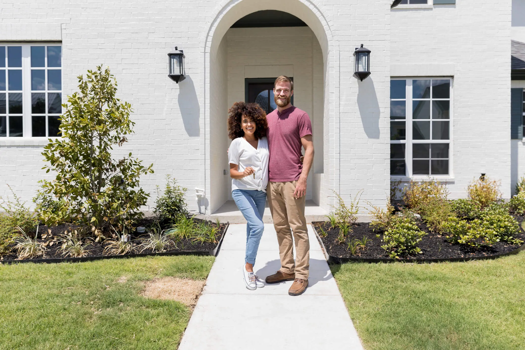 Excited expectant couple holds the keys to their new home. They are standing in front of the house on a sunny day.
