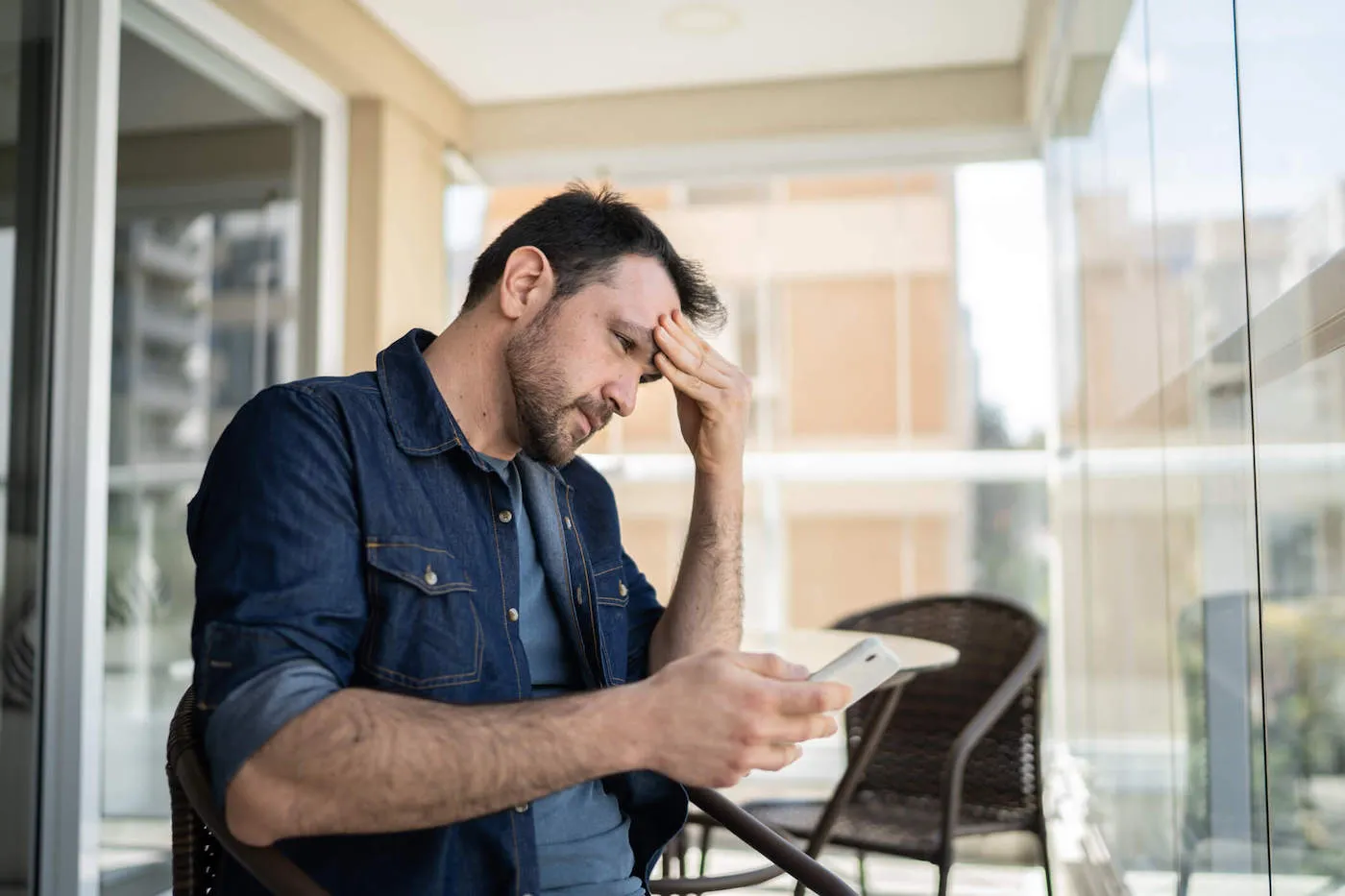 A man wearing a blue shirt frowns as he has his hand to his face while he looks at his phone.
