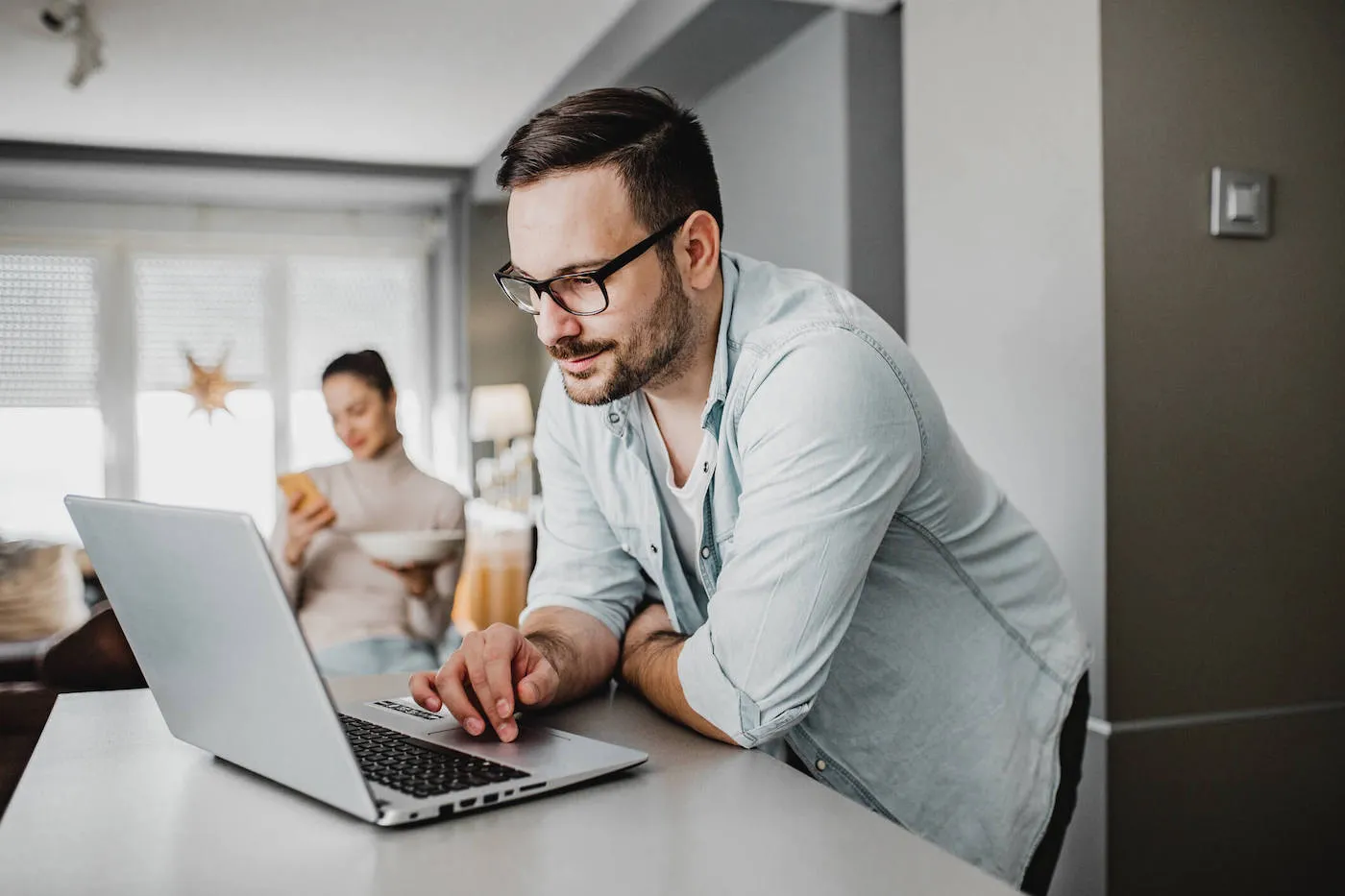 A man wearing a light blue shirt is looking at his laptop at the kitchen counter while a women uses her phone in the background.