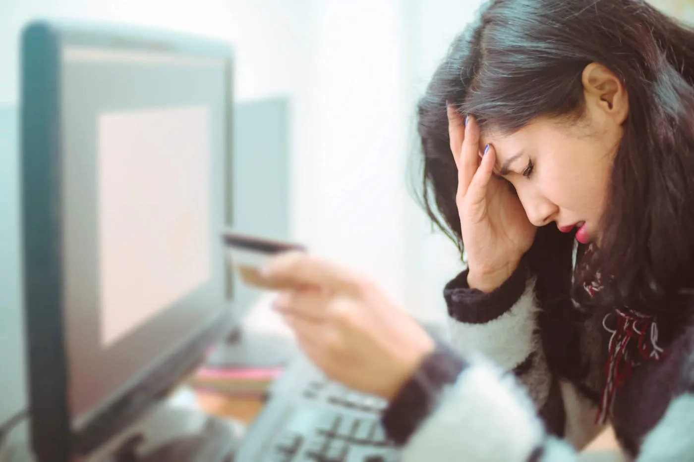 A woman has her hand to her face while holding a credit card in front of her computer.