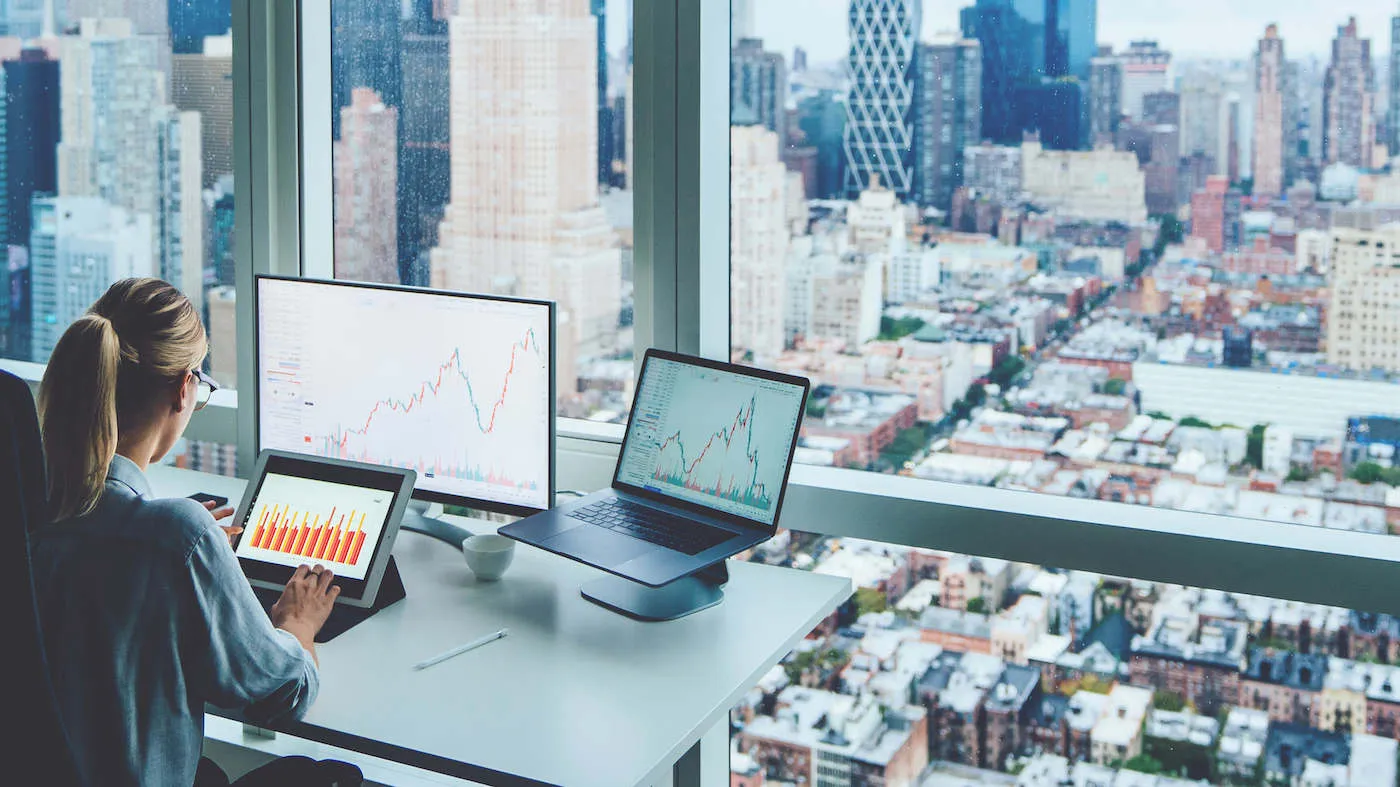 A woman sitting down uses her tablet while her two other computers are on her desk with the a city view from her window.