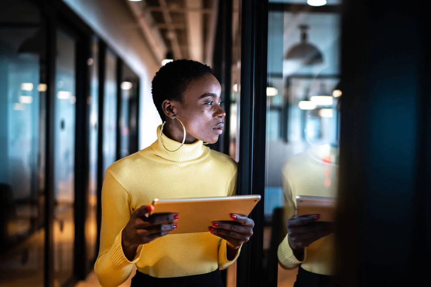 A woman wearing an orange turtleneck sweater holds a tablet while looking at a window screen at the office.
