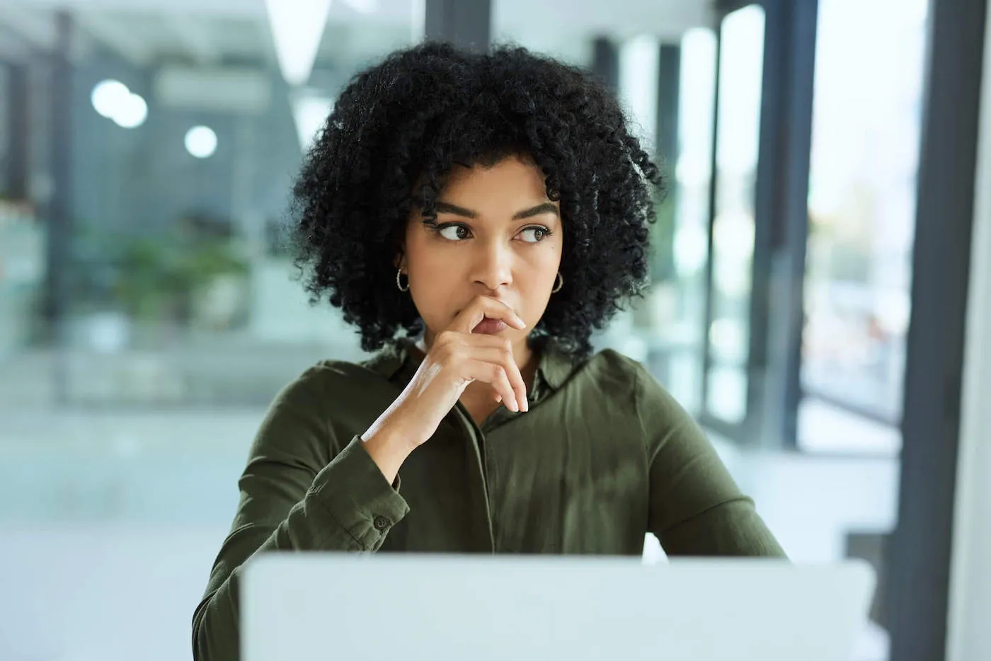 A young women with curly hair and a green sweater sits in front of her computer as she has her hand over her mouth while thinking.