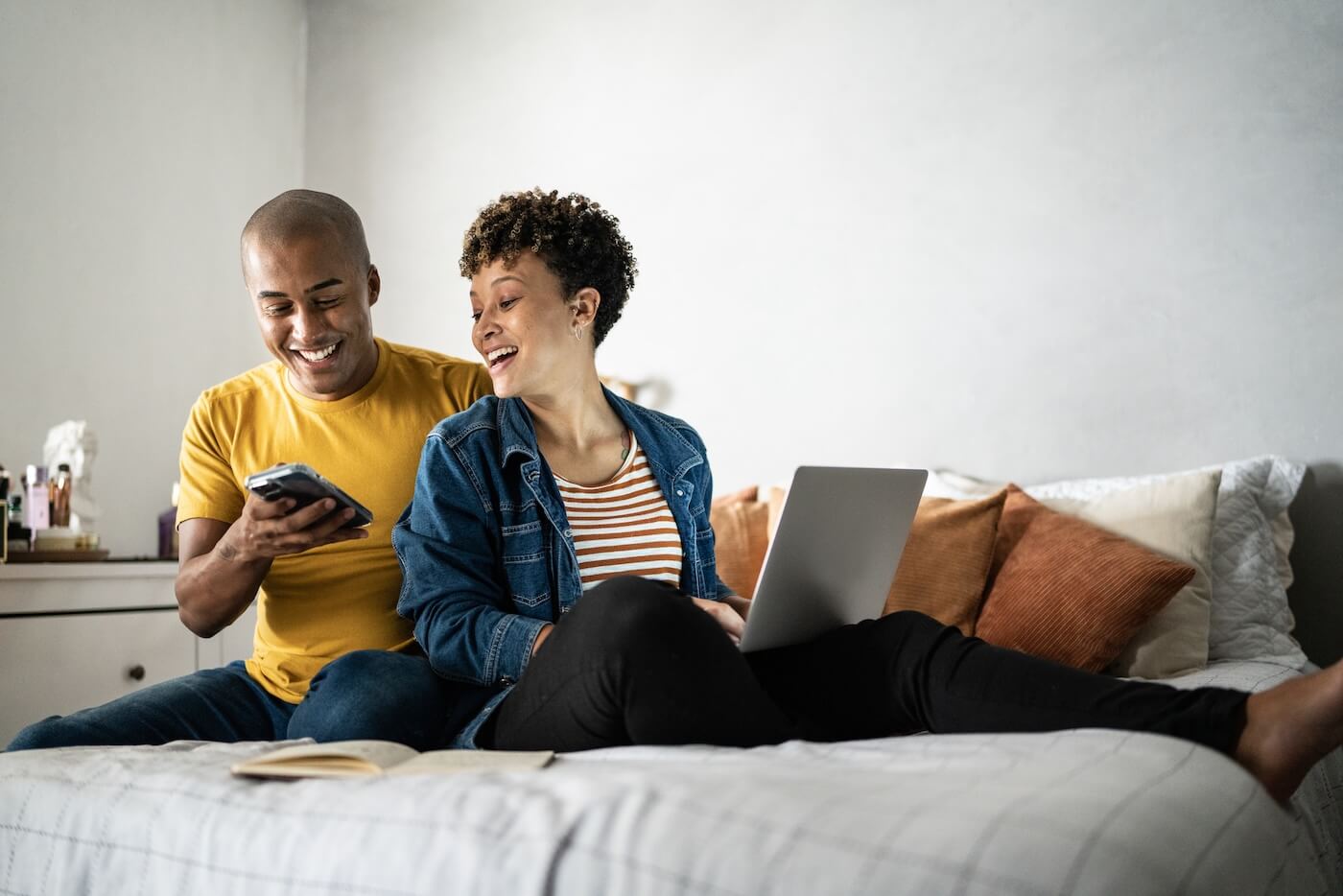 Smiling couple sitting on the bed with a laptop while checking a smartphone