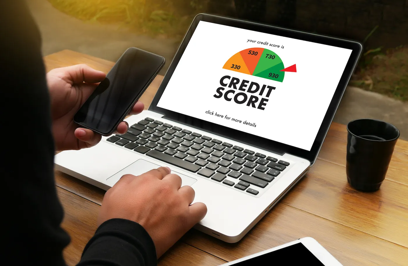 A person holding their mobile phone and checking their credit score on a laptop on their desk.