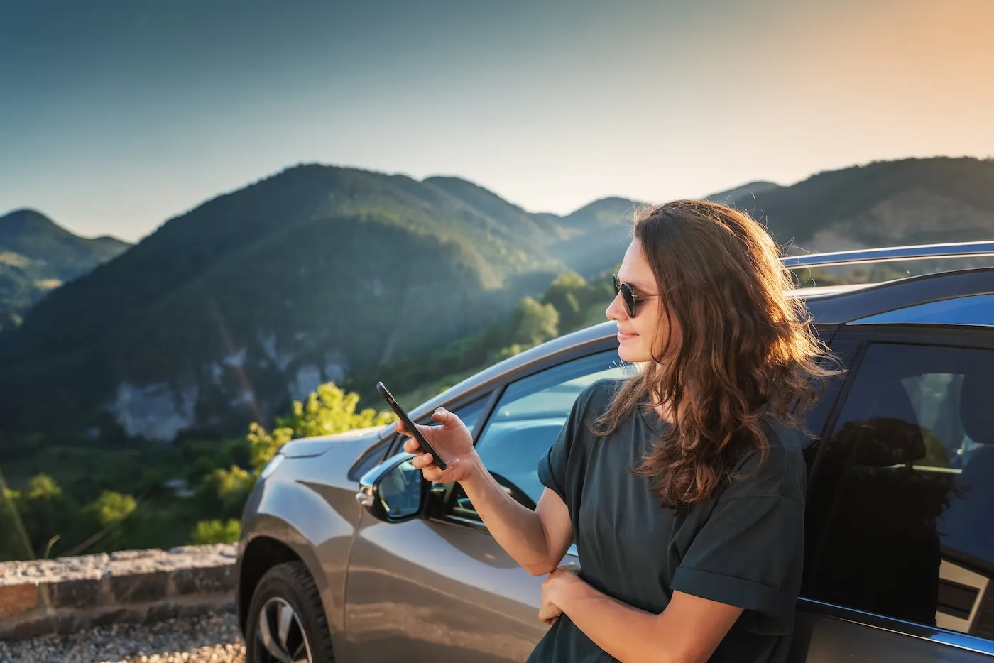 Young woman traveling by car in the mountains using smartphone at sunset.