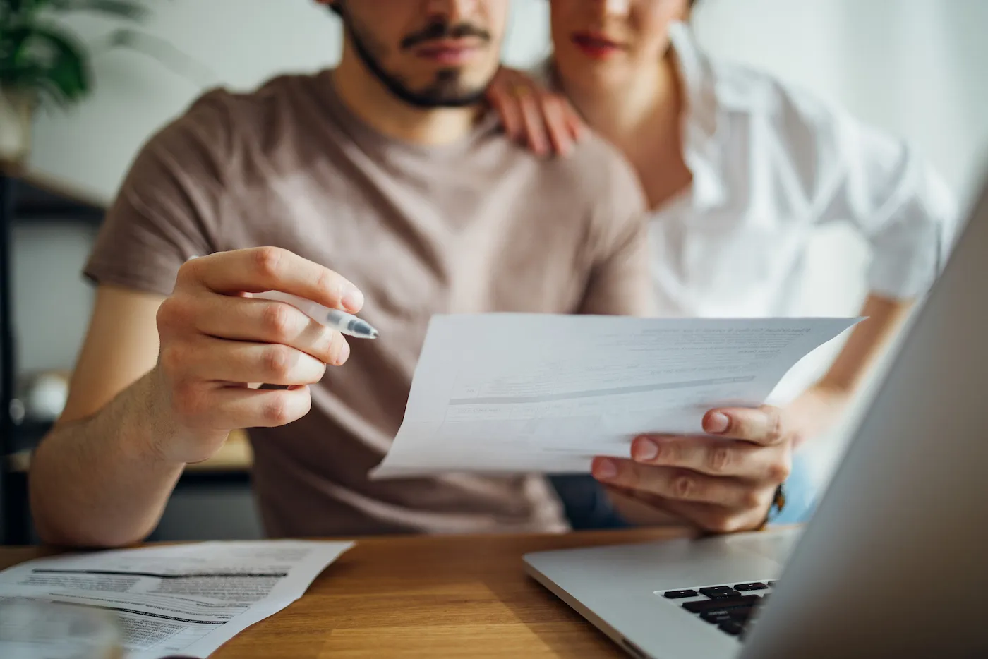 Close up photo of male hands holding bill and pen and female hands hugging him while they doing home finances together online on a laptop computer in the kitchen.