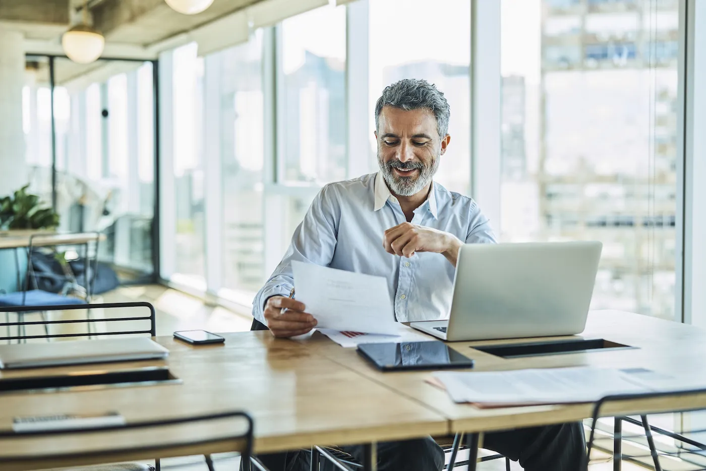 A smiling man checking his credit score, sitting at a desk next to a window and holding papers.