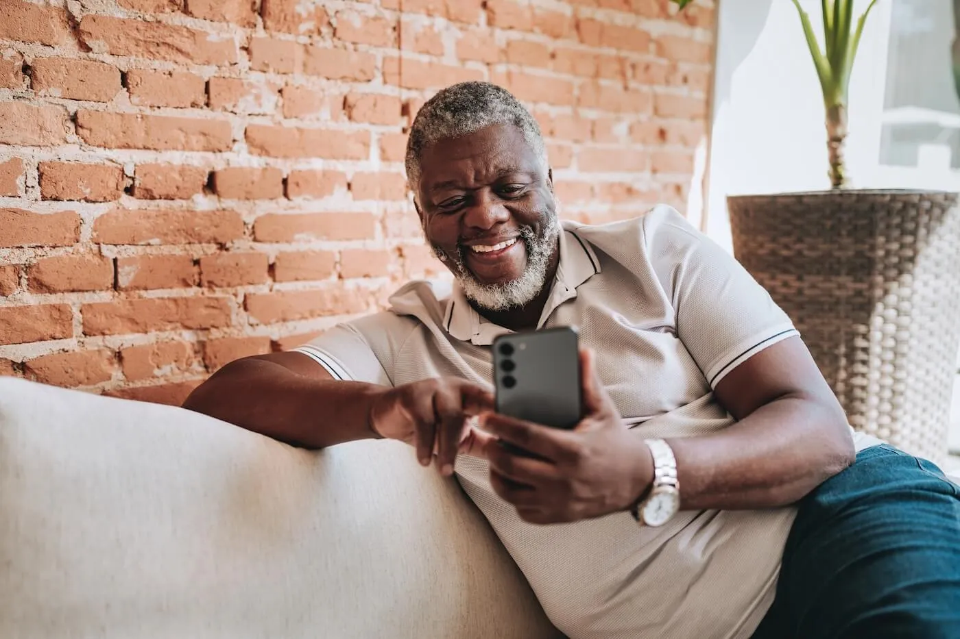 Smiling senior man sitting on a couch next to a brick wall and using his smartphone