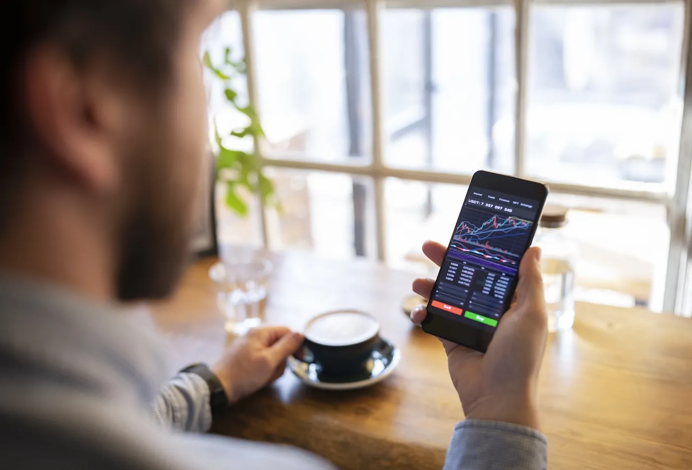Young unrecognisable man using investing app on smart phone and having coffee.