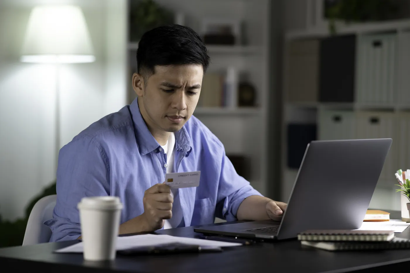 A worriedd man sitting in front of his laptop holding a credit card, considering canceling it after a data breach.